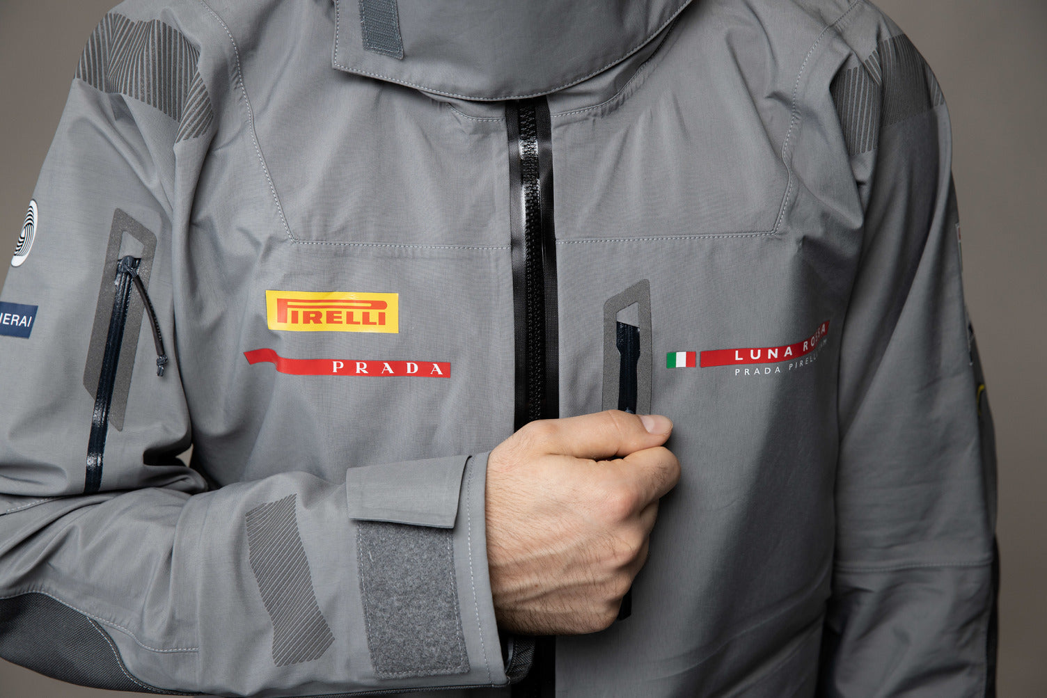 A close-up of a gray technical sailing jacket with a black zipper, featuring the Pirelli, Prada, and Luna Rossa logos on the chest.
