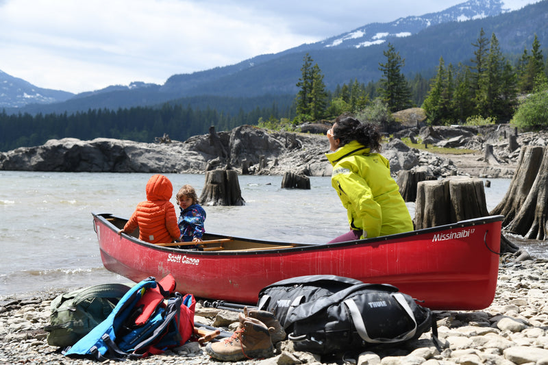 A woman and two young children sit in a red canoe on a rocky shore, with gear bags in the foreground and a mountain lake behind them.
