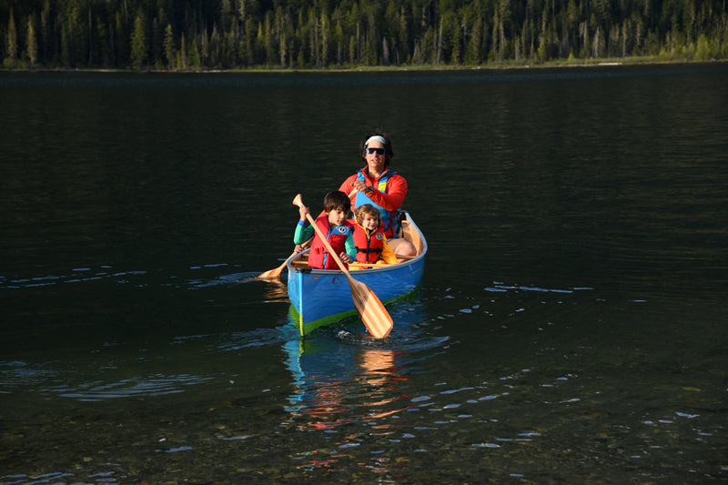 A father and his two young children, all wearing life jackets, paddle a blue canoe across a calm lake on a sunny day.