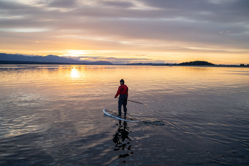 A person paddles a stand-up paddleboard on calm water during a golden sunset, with mountains silhouetted in the distance.