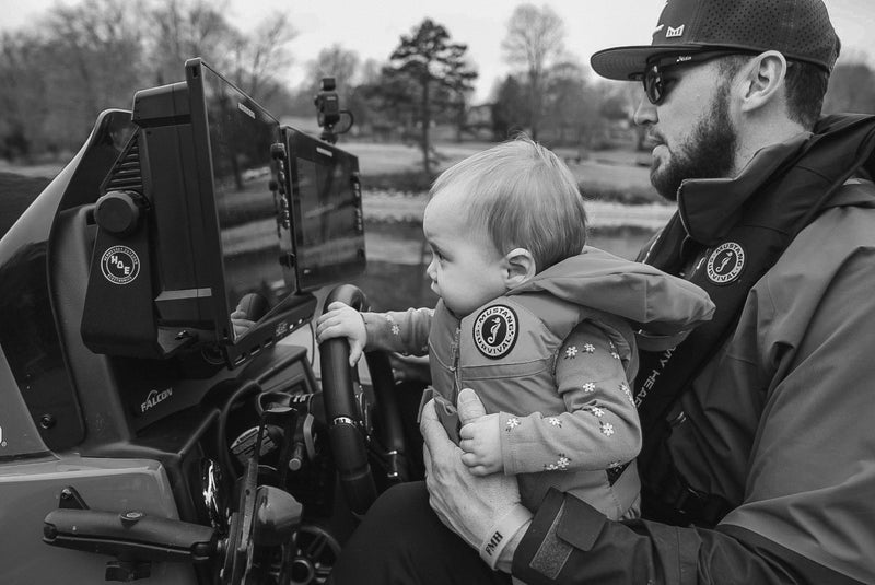 Father holding a content child on his lap while driving a boat. She's wearing an infant Lil Legends PFD.