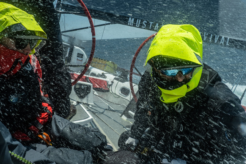Two sailors on a racing yacht endure heavy ocean spray while wearing high-visibility foul weather gear and hoods.