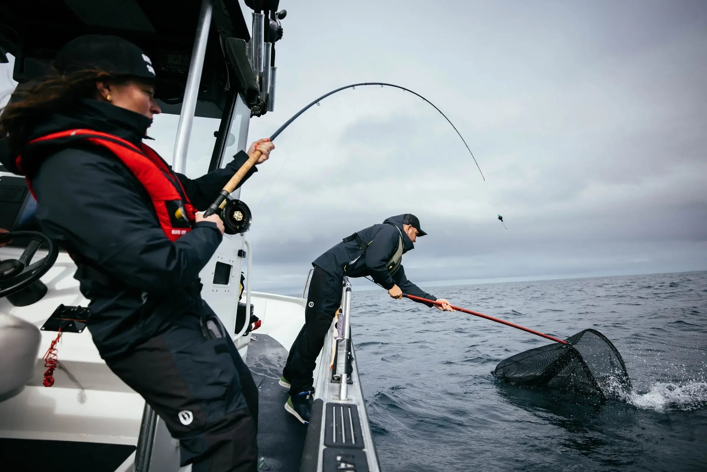 Anglers working together on a boat as one reels in a fish and the other prepares a landing net.