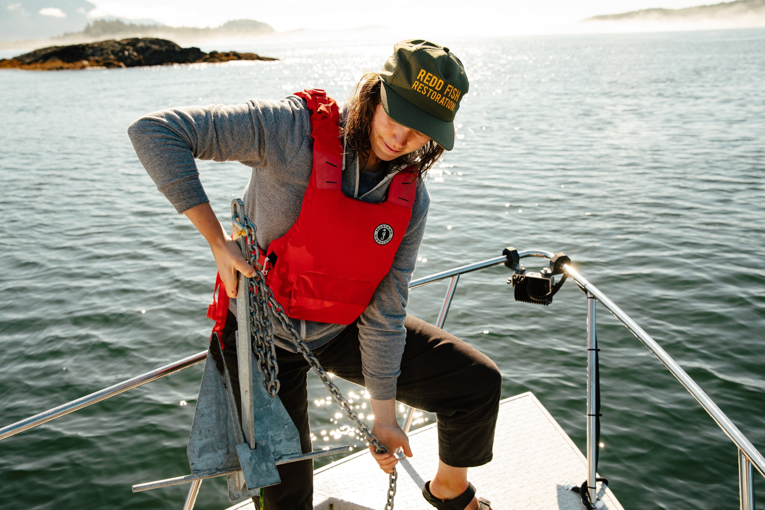 A Redd Fish Restoration crew member lifts a heavy metal anchor and chain while wearing a red Khimera flotation vest.