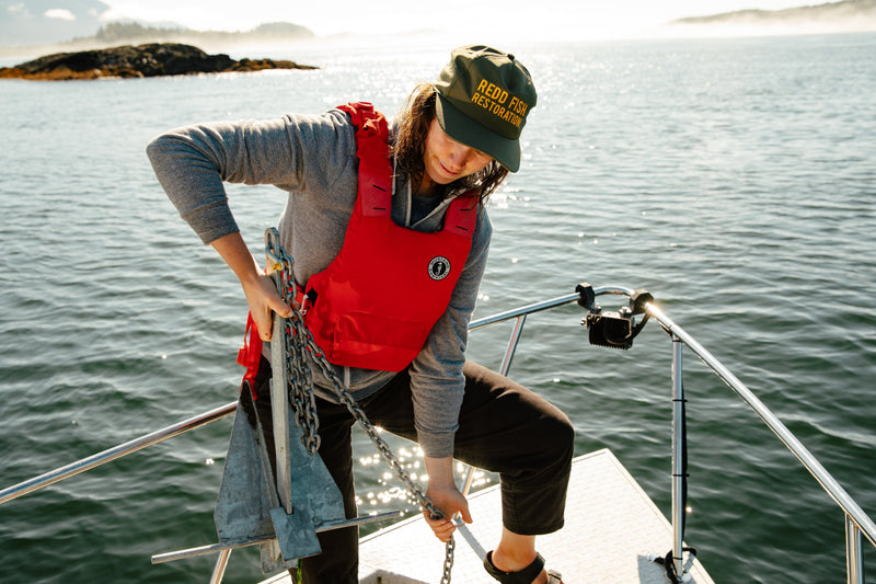 A Redd Fish Restoration crew member lifts a heavy metal anchor and chain while wearing a red Khimera flotation vest.