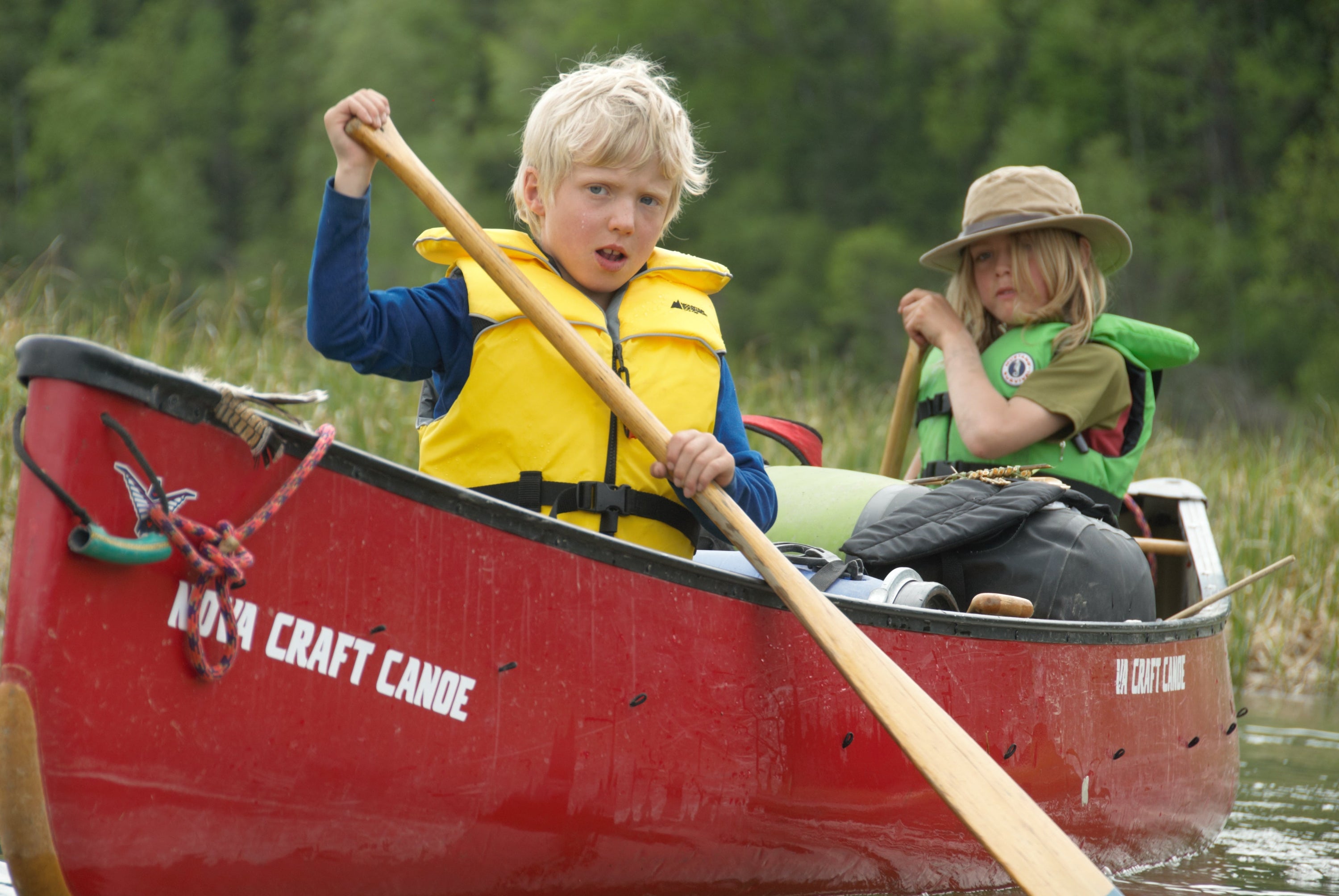 A blonde boy and another child wearing kids life jackets paddle a red Nova Craft Canoe together near a grassy shoreline.