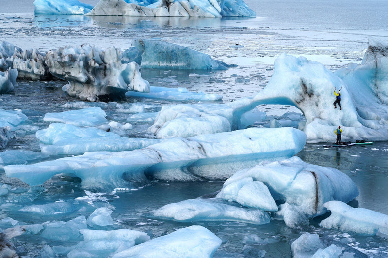 A person climbs a massive blue iceberg with ice axes while another person watches from a paddleboard in an icy field of smaller formations.