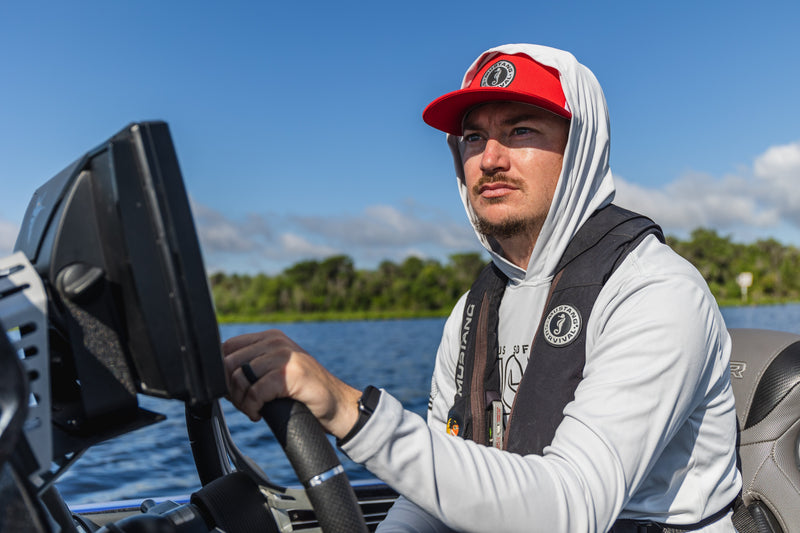 Angler Hunter Bland wears a red cap, white hoodie, and black life vest while steering his boat on a sunny day.