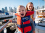 Child wearing a life jacket with a woman on a boat in a city harbor.