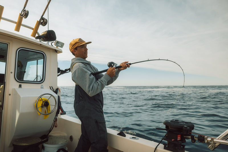Brody Pigeau wears a yellow cap and Taku waterproof bibs while reeling in a big fish from the deck of his boat.