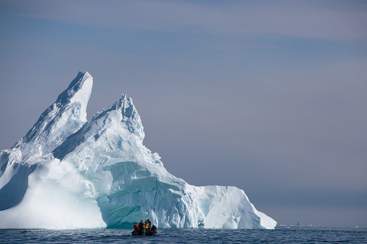 A small zodiac boat with travelers floats several hundred meters away from a massive, towering iceberg near Vernadsky Station.