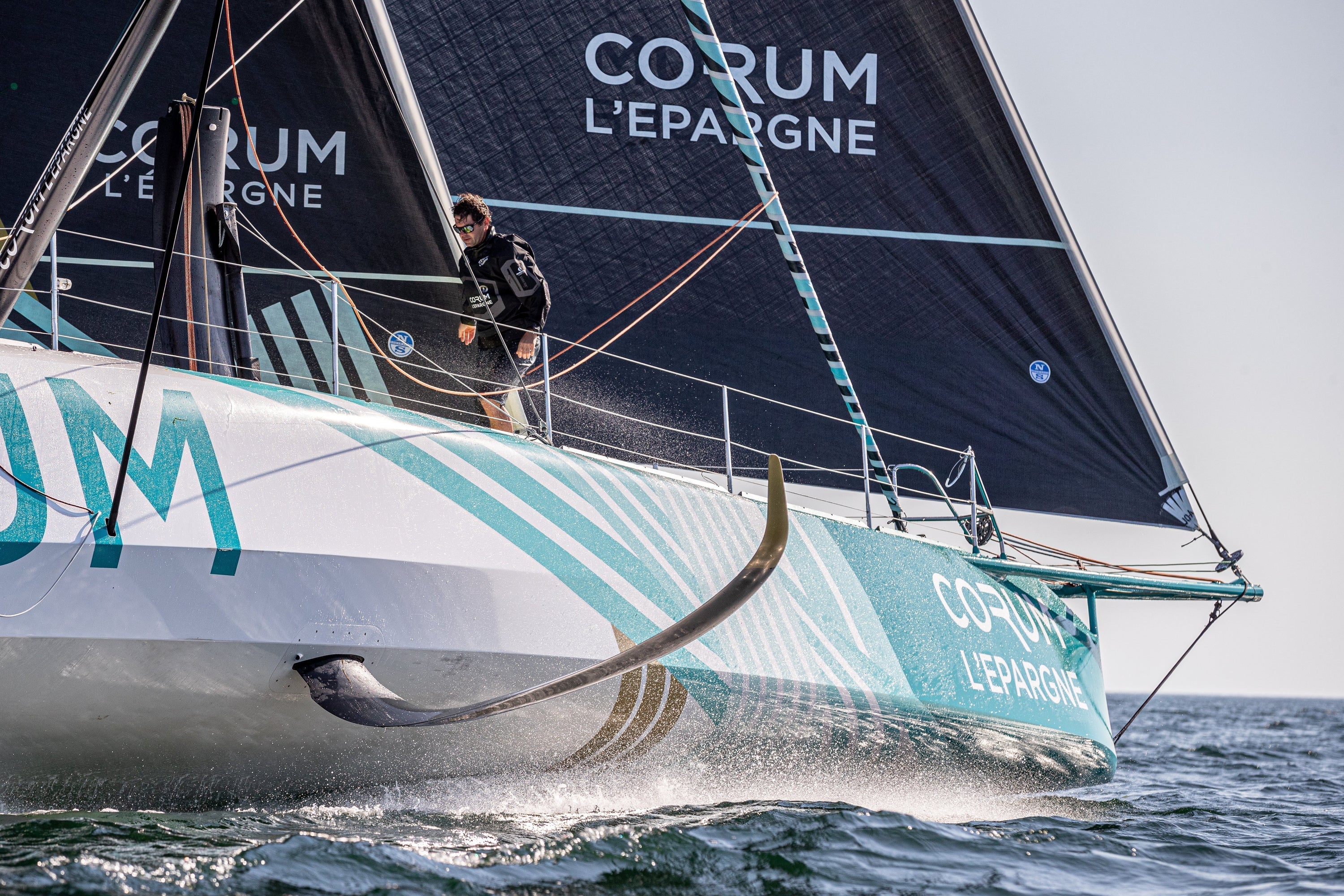 Skipper Nicolas Troussel stands on the deck of the CORUM L'Épargne IMOCA sailboat as it slices through the ocean.