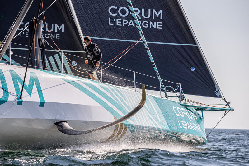 Skipper Nicolas Troussel stands on the deck of the CORUM L'Épargne IMOCA sailboat as it slices through the ocean.