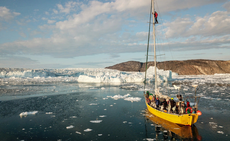 A yellow sailboat navigates an icy sea near a massive glacier, while a crew member stands high up on the mast to scout the path ahead.