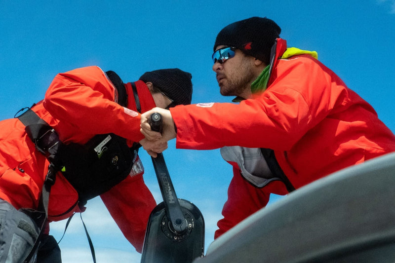 Two crew members wearing Meris gear and Elite 28 hydrostatic PFDs work together on a sailboat winch pedestal.