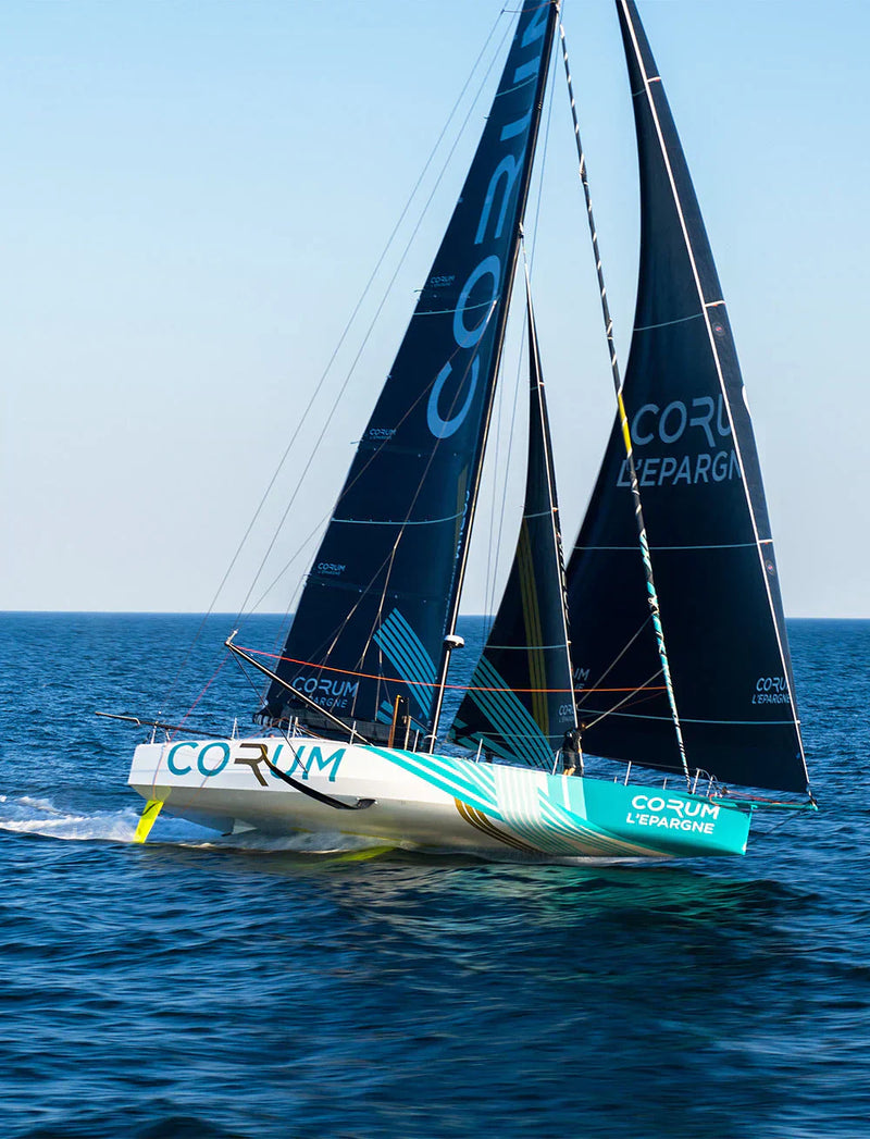 The blue and white racing yacht Corum L'Épargne speeds through the ocean with its large black sails fully deployed.