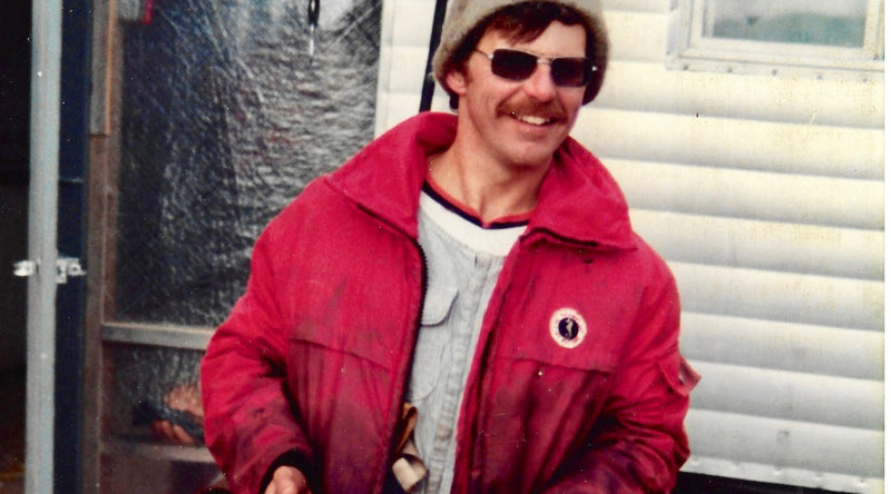 A man with a mustache wearing a beanie, sunglasses, and a red Mustang flotation coat smiles in front of a white trailer.
