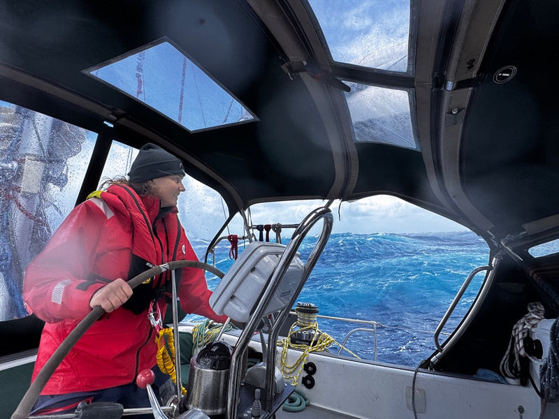 A sailor in a red Meris waterproof jacket steers a sailboat from a covered helm station as massive waves churn behind.