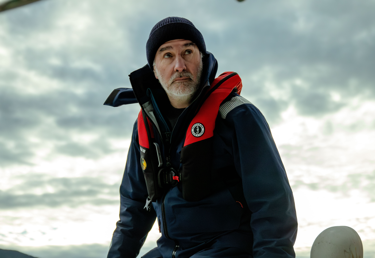 A sailor wearing a black beanie, a navy jacket, and a red Mustang Survival Atlas life jacket looks upward against a cloudy sky.