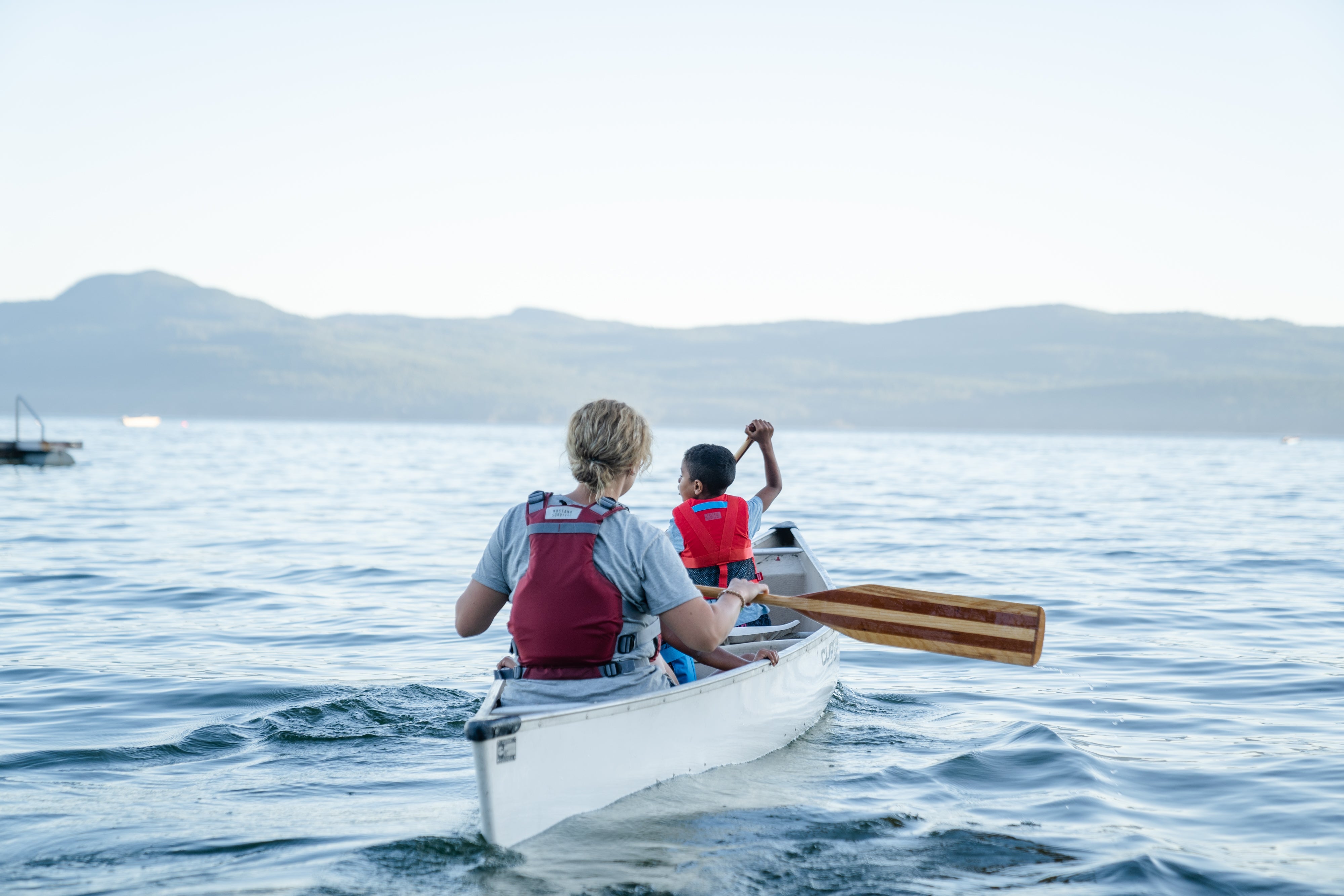 Two paddlers wearing red life jackets canoe across calm water with a mountain landscape in the background