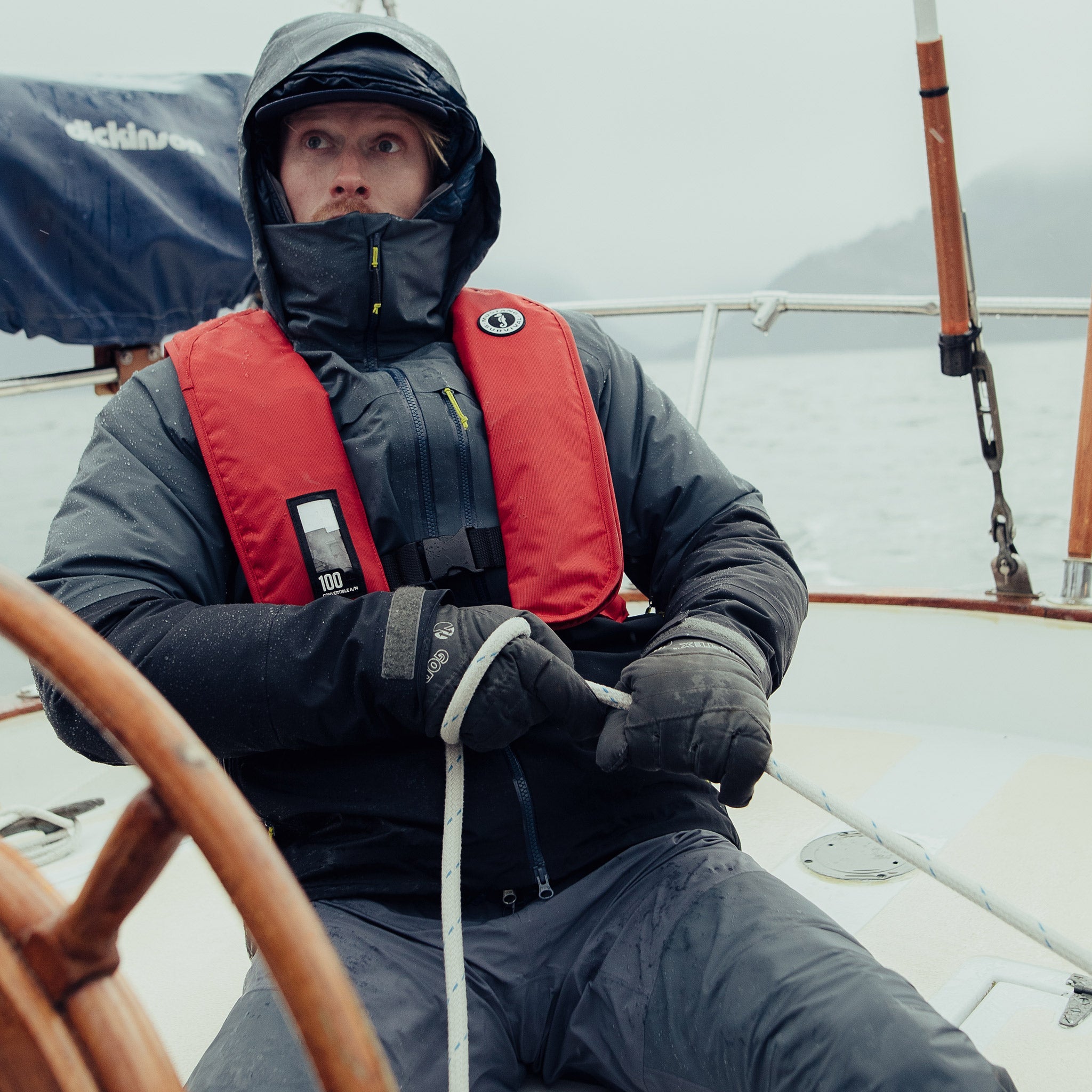 A sailor pulling a line on a sailboat deck while wearing a dark hooded foul-weather jacket and a red Mustang Survival MIT 100 Convertible inflatable PFD.