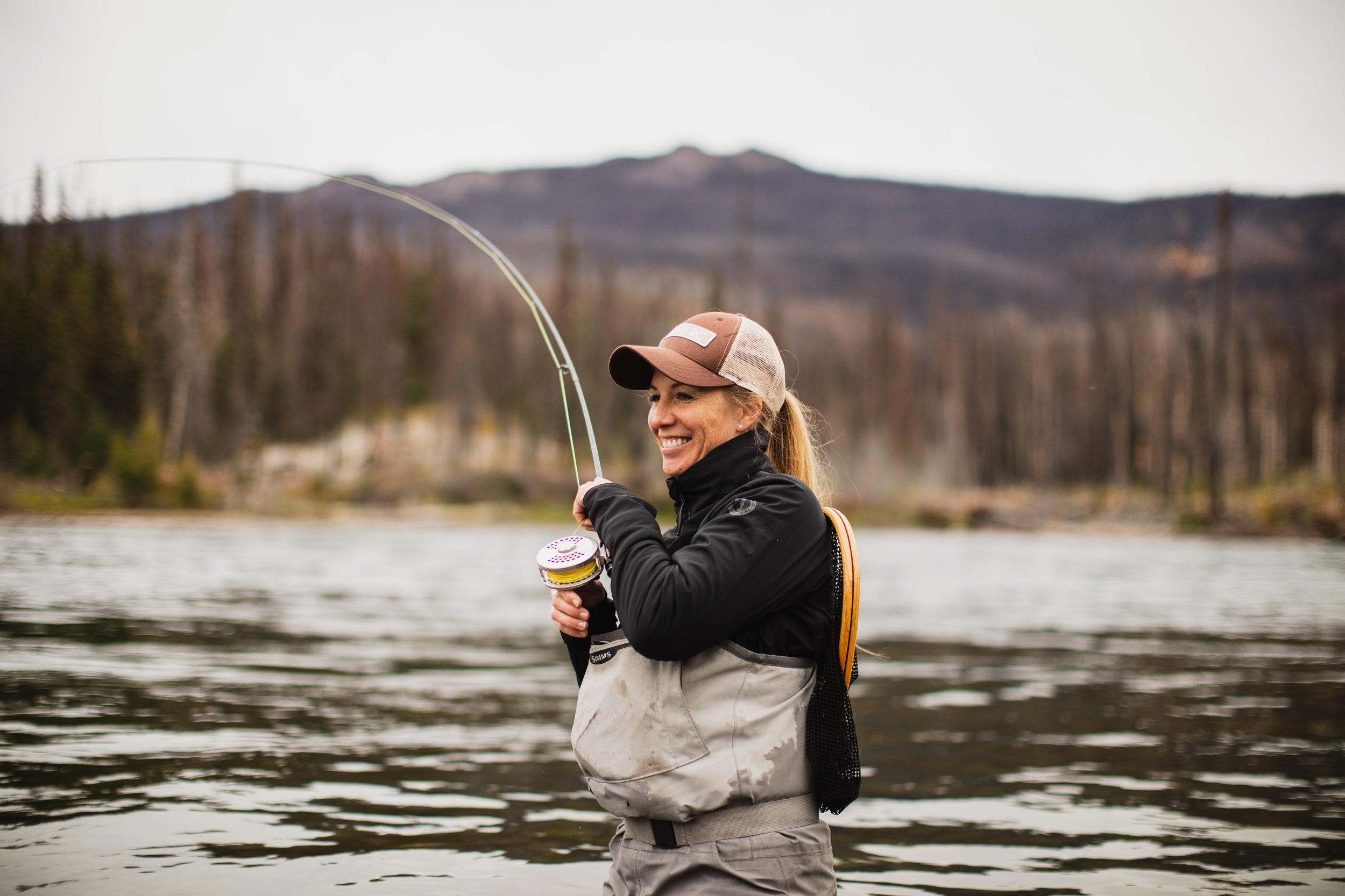 Woman wearing Mustang Survival gear, smiling while river fishing with a bent rod, standing in shallow water surrounded by forest and mountains.