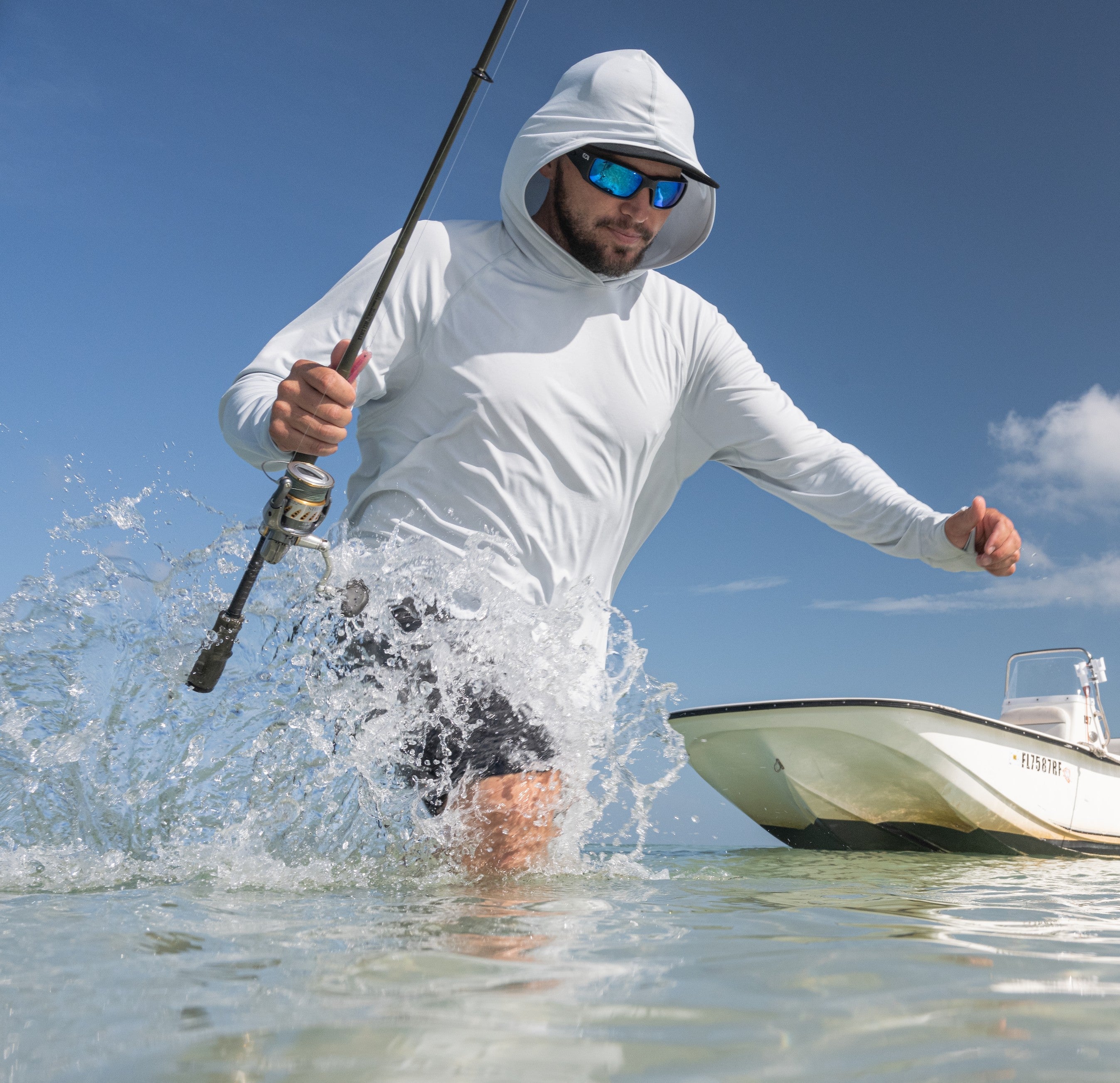 An angler wading through ocean water holding a fishing rod next to a skiff boat, wearing a white Mustang Survival long sleeve UV hoodie and polarized sunglasses.