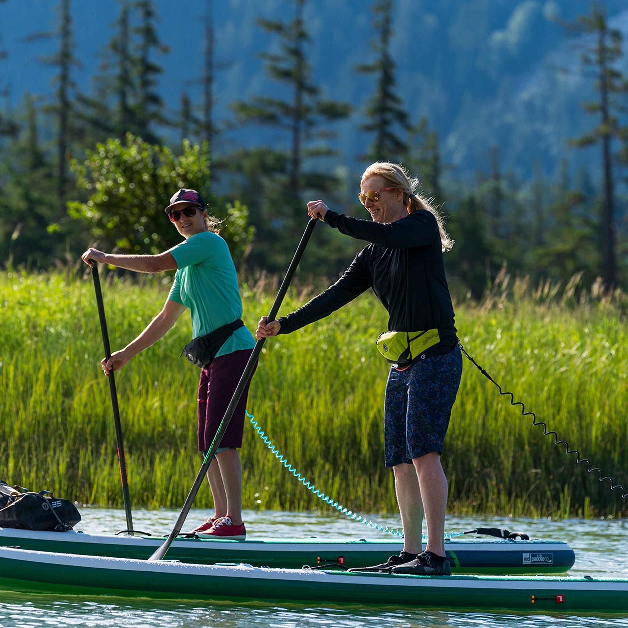 Two women stand-up paddleboarding on a calm river while wearing low-profile Mustang Survival inflatable belt pack PFDs around their waists.
