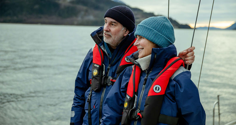 Two people in blue jackets with red life vests standing on a boat with a scenic background.
