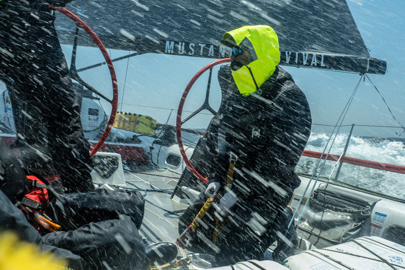Person on a sailboat in rough weather wearing a bright yellow hood and dark clothing.